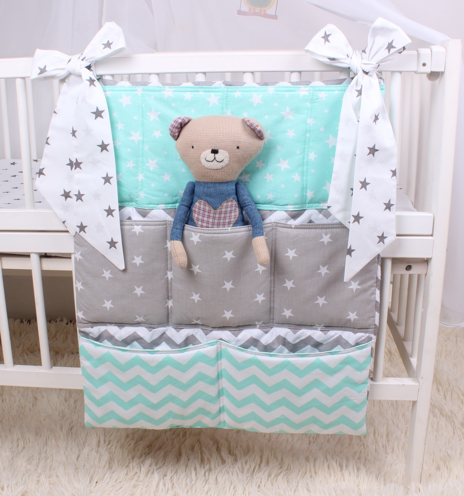Organizer na dziecięce akcesoria w biało-turkusowo-szare gwiazdki, zawieszony na białym łóżeczku, z pluszowym misiem w jednej z kieszeni.