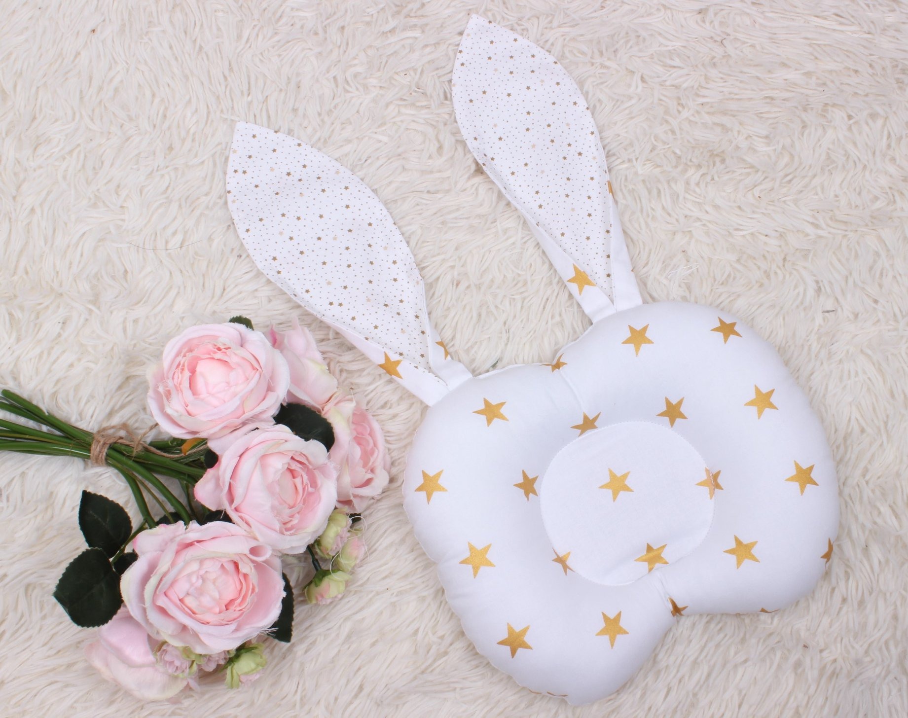 Biała poduszka dla niemowląt w kształcie króliczka z złotymi gwiazdkami, leżąca na puszystym białym dywanie obok bukietu bladoróżowych róż.