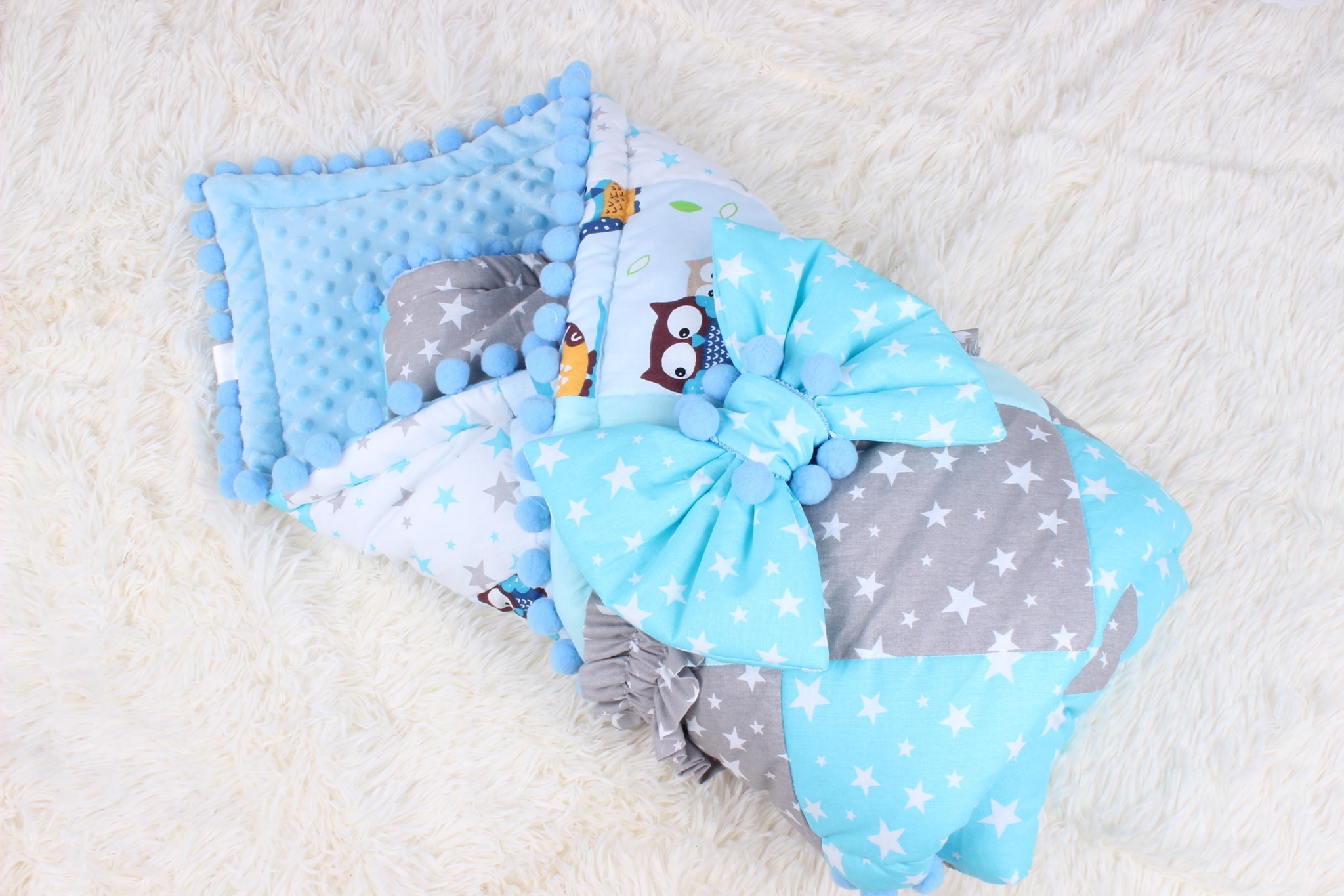 Komplet niemowlęcy: pikowana kołderka z motywem sowy i gwiazd oraz poduszka z wypustkami, wykończone niebieskimi pomponami, na białym, puszystym tle.