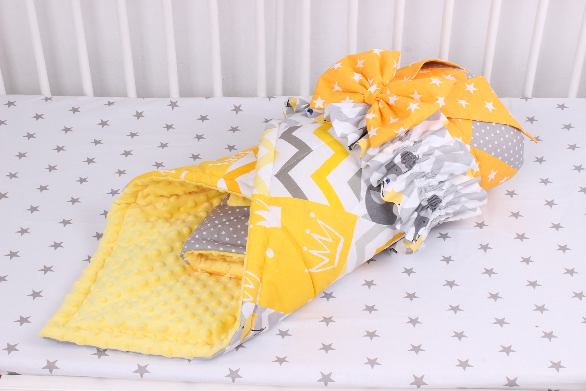 Komplet pościeli dziecięcej w łóżeczku: żółty kocyk z wypukłą fakturą, poduszka z motywem korony i zygzaków, oraz poduszka z motywem słoni, całość w odcieniach żółci, szarości i bieli.