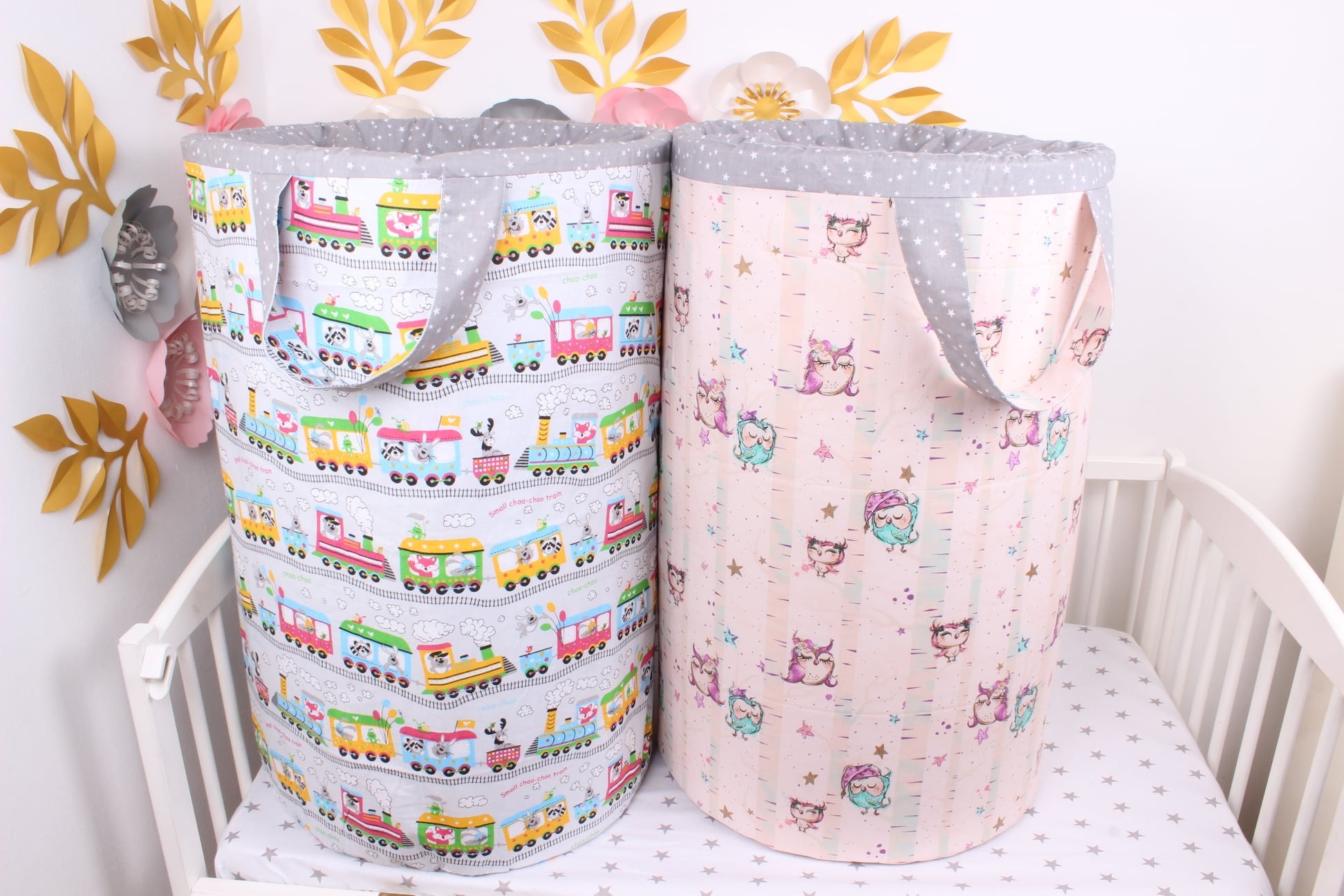 Dwa kosze na zabawki lub pranie dziecięce, jeden z motywem pociągów ze zwierzętami, drugi z motywem śpiących sów, oba z szarym obszyciem w białe gwiazdki, stojące w białym łóżeczku dziecięcym.