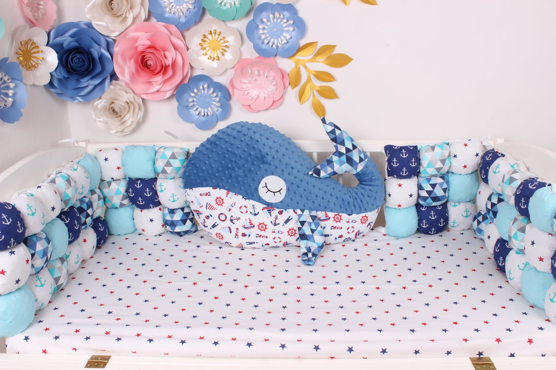 Dziecięce łóżeczko z motywem morskim: niebieski pluszowy wieloryb, ochraniacze w marynarskie wzory (kotwice, trójkąty), pościel w czerwone i niebieskie gwiazdki, dekoracje z papierowych kwiatów...
