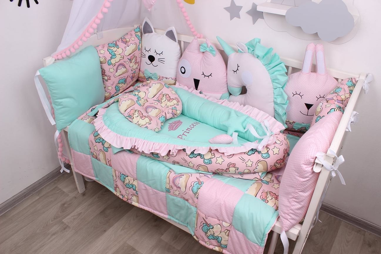 Łóżeczko dziecięce z patchworkową kołderką w pastelowych kolorach, poduszkami w kształcie zwierząt i kokonem niemowlęcym z napisem 'Princess'.