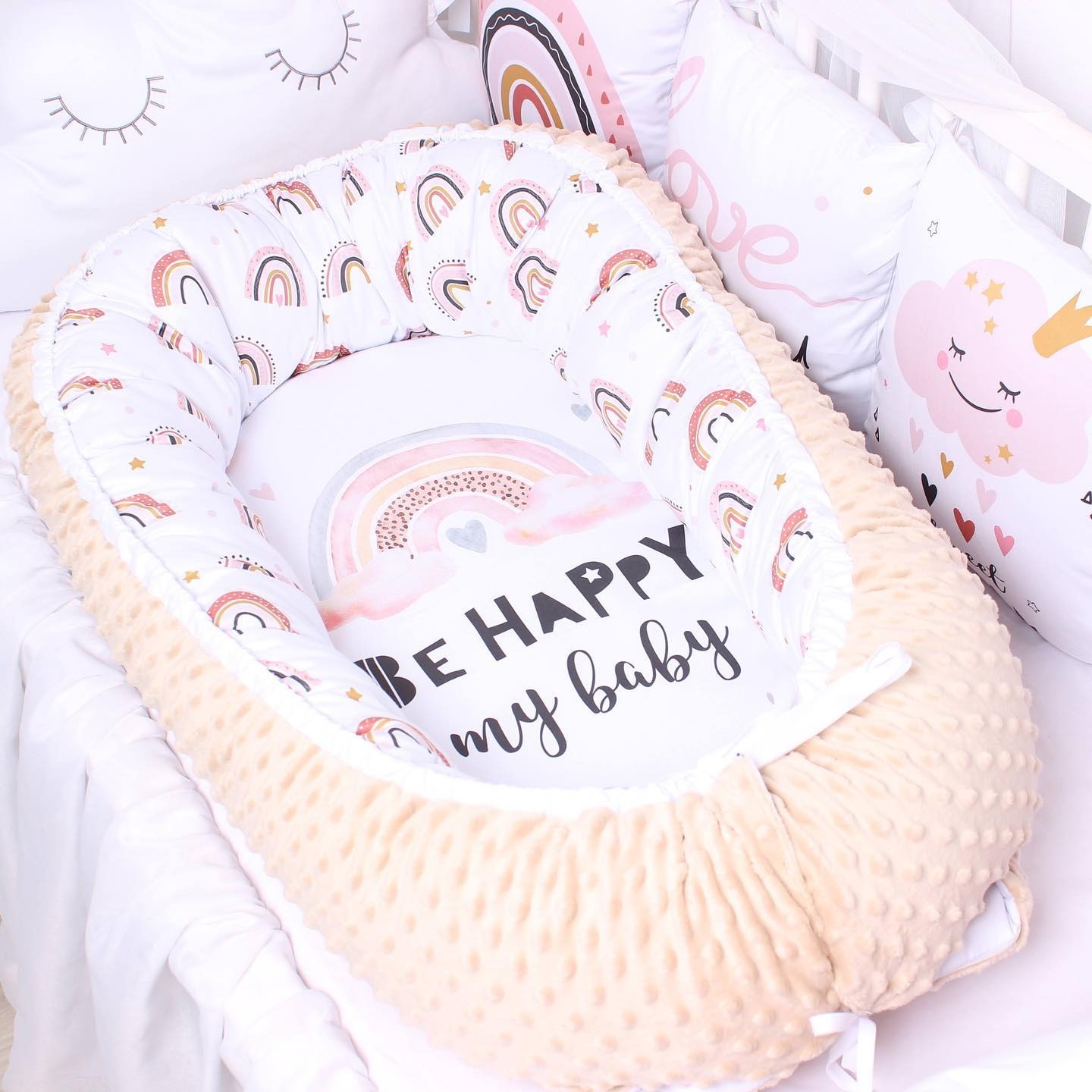 Beżowe gniazdko niemowlęce z motywem tęczy i napisem 'Be Happy my baby', otoczone poduszkami z motywami chmurek i tęczy w łóżeczku.