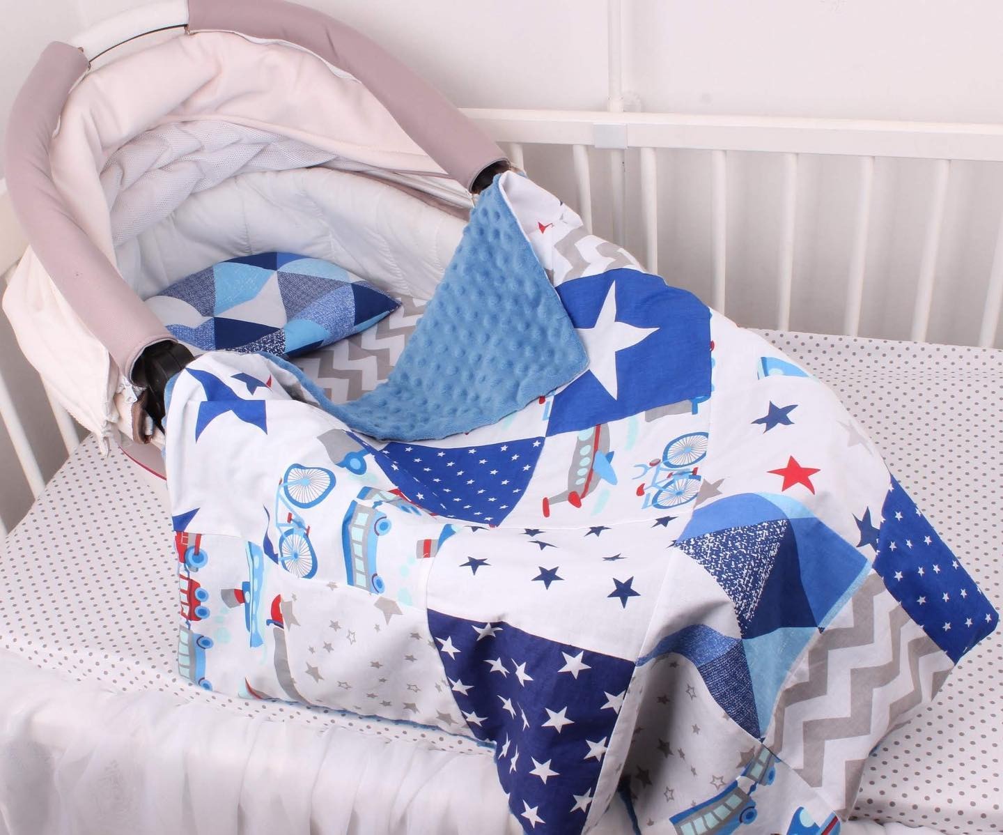 Kołderka patchworkowa w kolorach niebieskim, białym i szarym z motywami gwiazd, samolotów i rowerów, leżąca w dziecięcym łóżeczku z poduszką w geometryczne wzory.