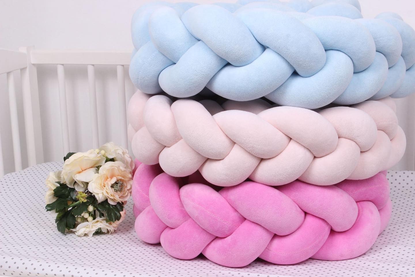 Trzy plecione ochraniacze do łóżeczka dziecięcego ułożone jeden na drugim: niebieski, różowy i beżowy, obok bukiet sztucznych kwiatów na pościeli w kropki.