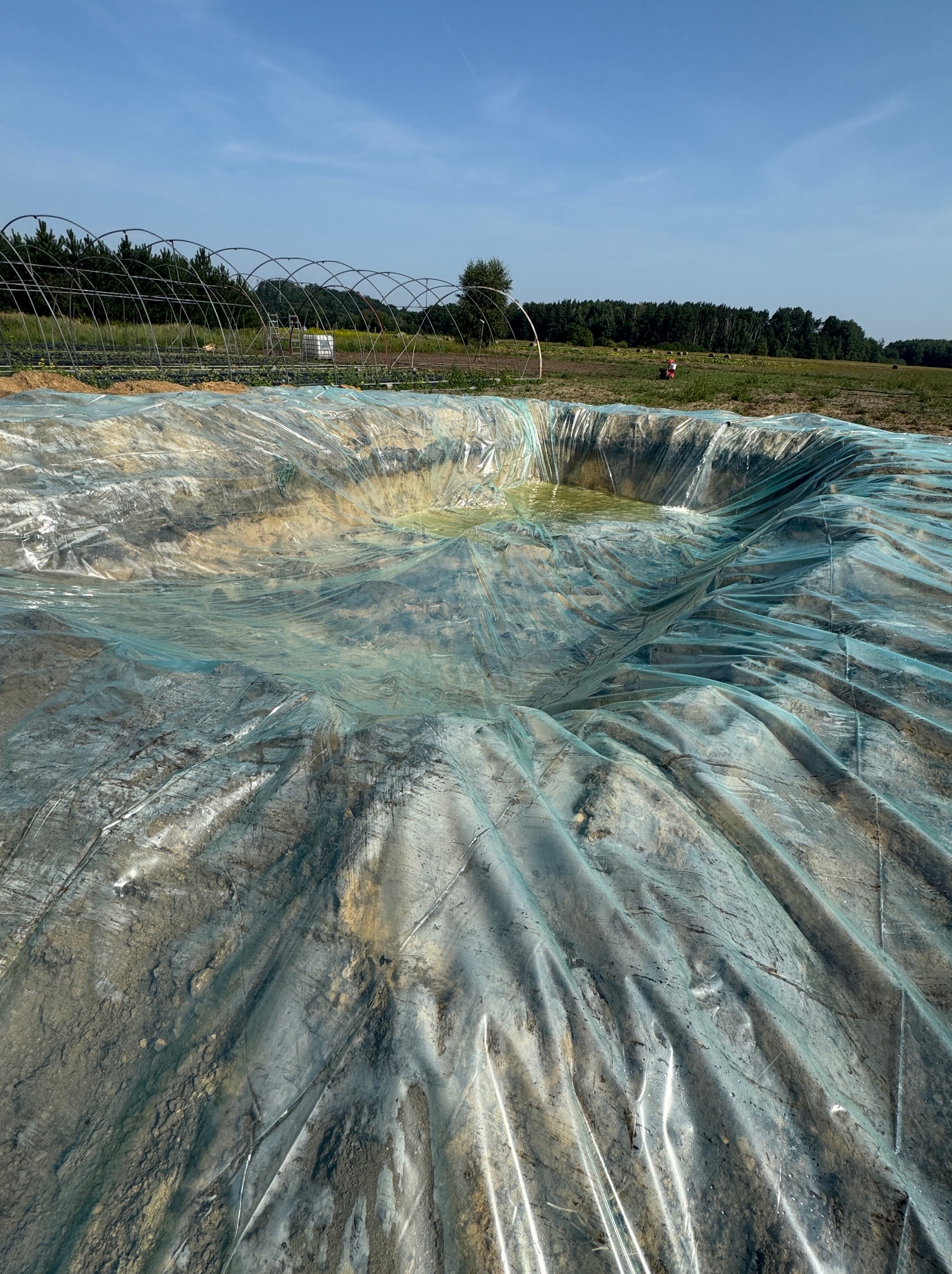Wykop ziemny zabezpieczony folią, w tle szklarnia i zieleń. Wykop częściowo wypełniony wodą. Ujęcie z góry, w słoneczny dzień.
