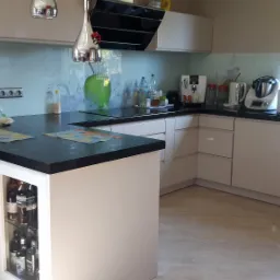 Jasna kuchnia z czarnym blatem i szafką z przeszklonym frontem, w której eksponowane są butelki alkoholu, z oknem w tle.