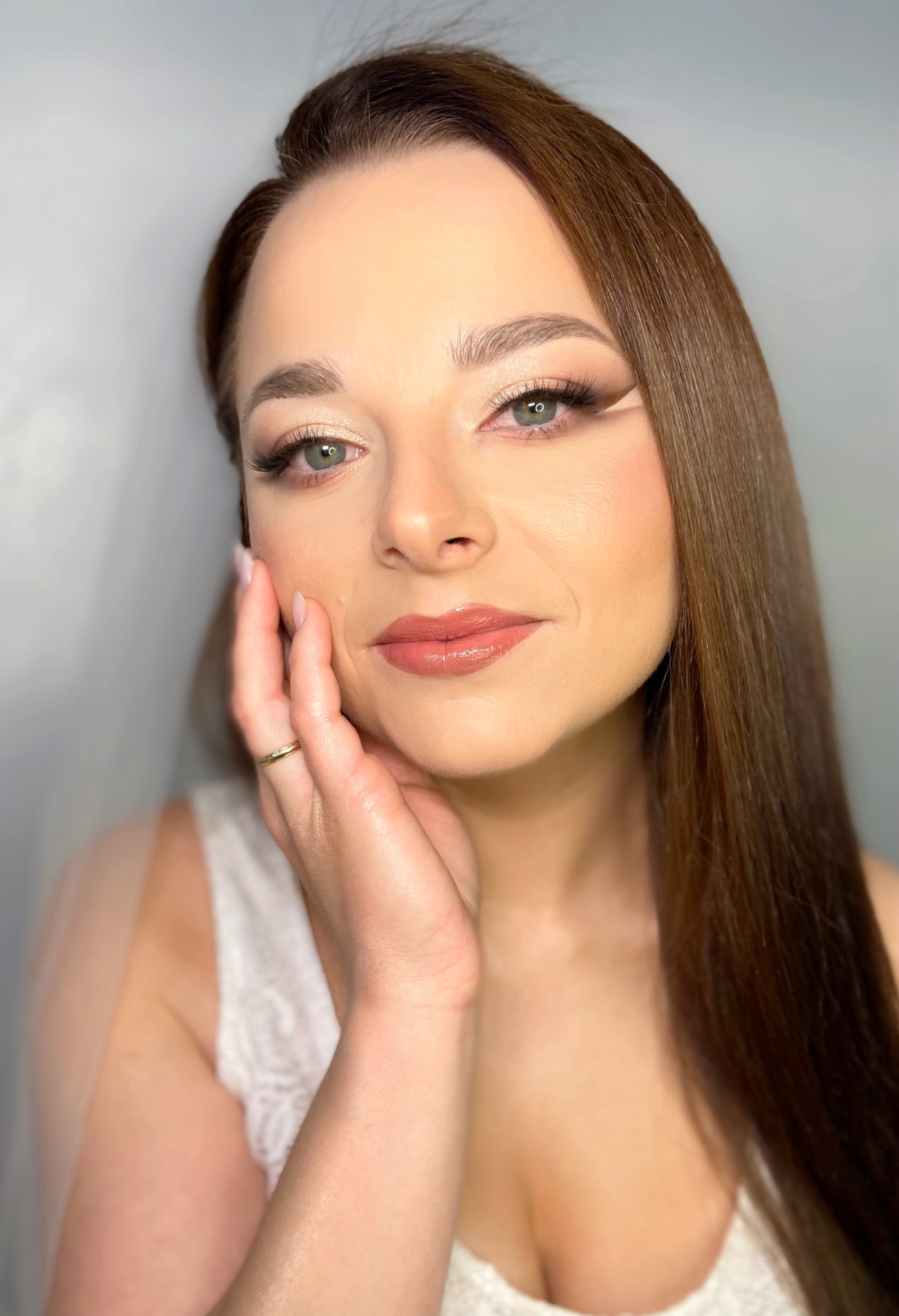 Portret kobiety z profesjonalnym, delikatnym makijażem oczu z eyelinerem i błyszczącymi ustami, dłoń z pierścionkiem dotyka policzka, subtelna suknia ślubna w tle.