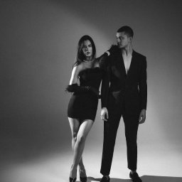 FOTOABERRACJA KAROL GACKOWSKI - Czarno-białe, studyjne ujęcie eleganckiej pary: kobieta w małej czarnej z piórami i perłami, mężczyzna w rozpiętym garniturze, oboje patrzą w bok.