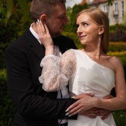FOTOABERRACJA KAROL GACKOWSKI - Uśmiechnięta para w plenerze: on w czarnym garniturze, ona w białej sukni z odkrytym ramieniem i z błyszczącym rękawem. W tle budynek i zieleń.