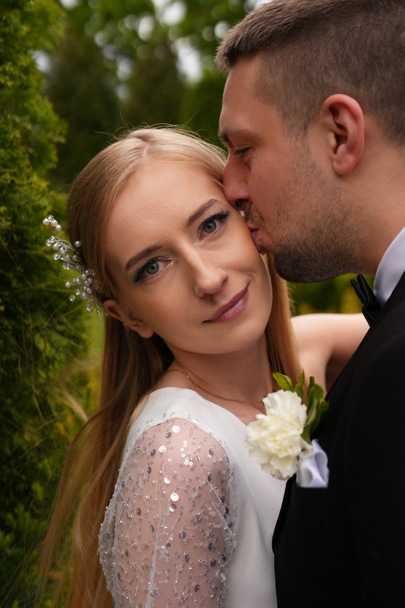 Uśmiechnięta panna młoda z ozdobą we włosach, całowana w policzek przez pana młodego w czarnym smokingu z butonierką na tle zieleni.