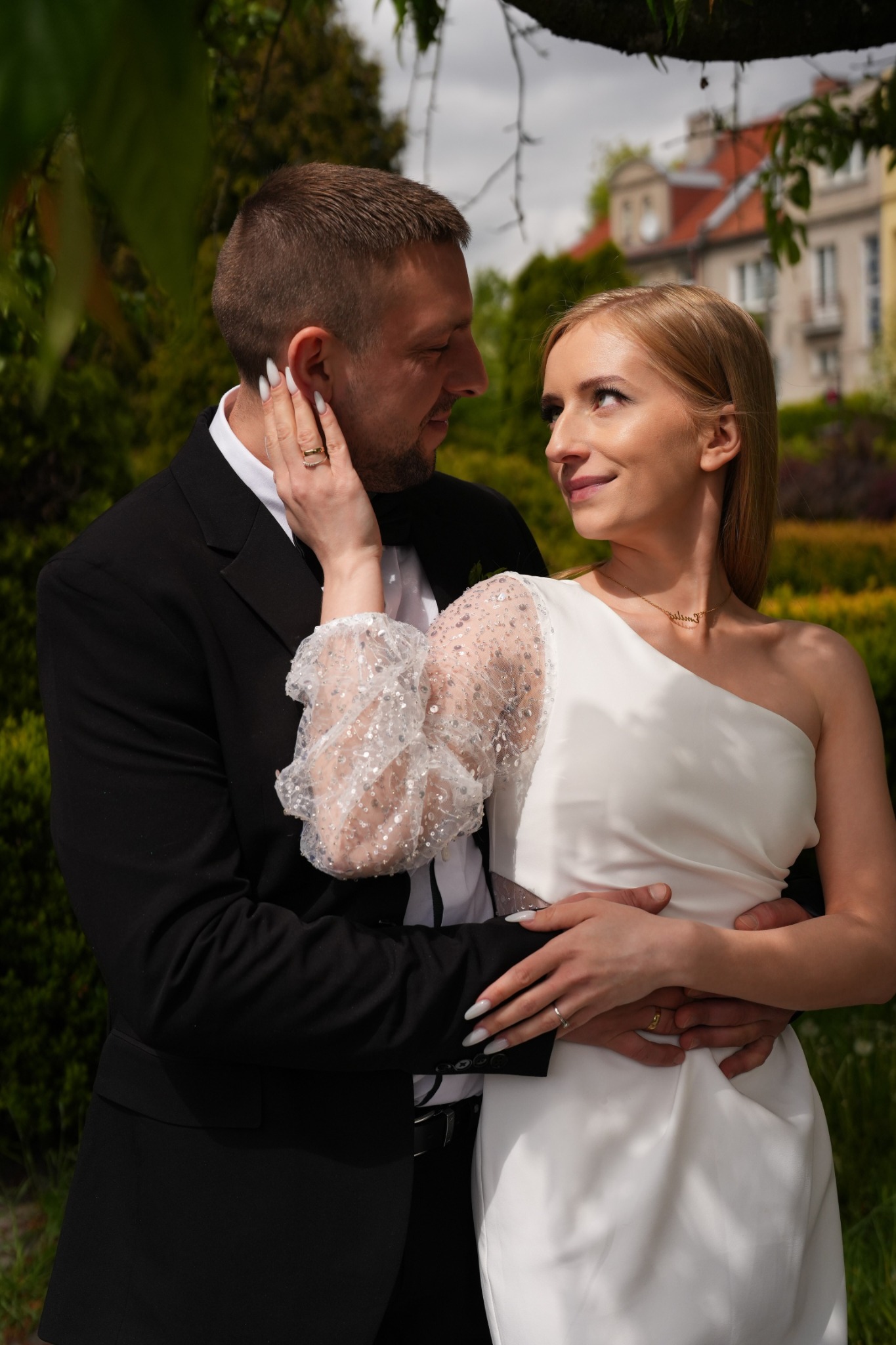 Uśmiechnięta para w plenerze: on w czarnym garniturze, ona w białej sukni z odkrytym ramieniem i z błyszczącym rękawem. W tle budynek i zieleń.