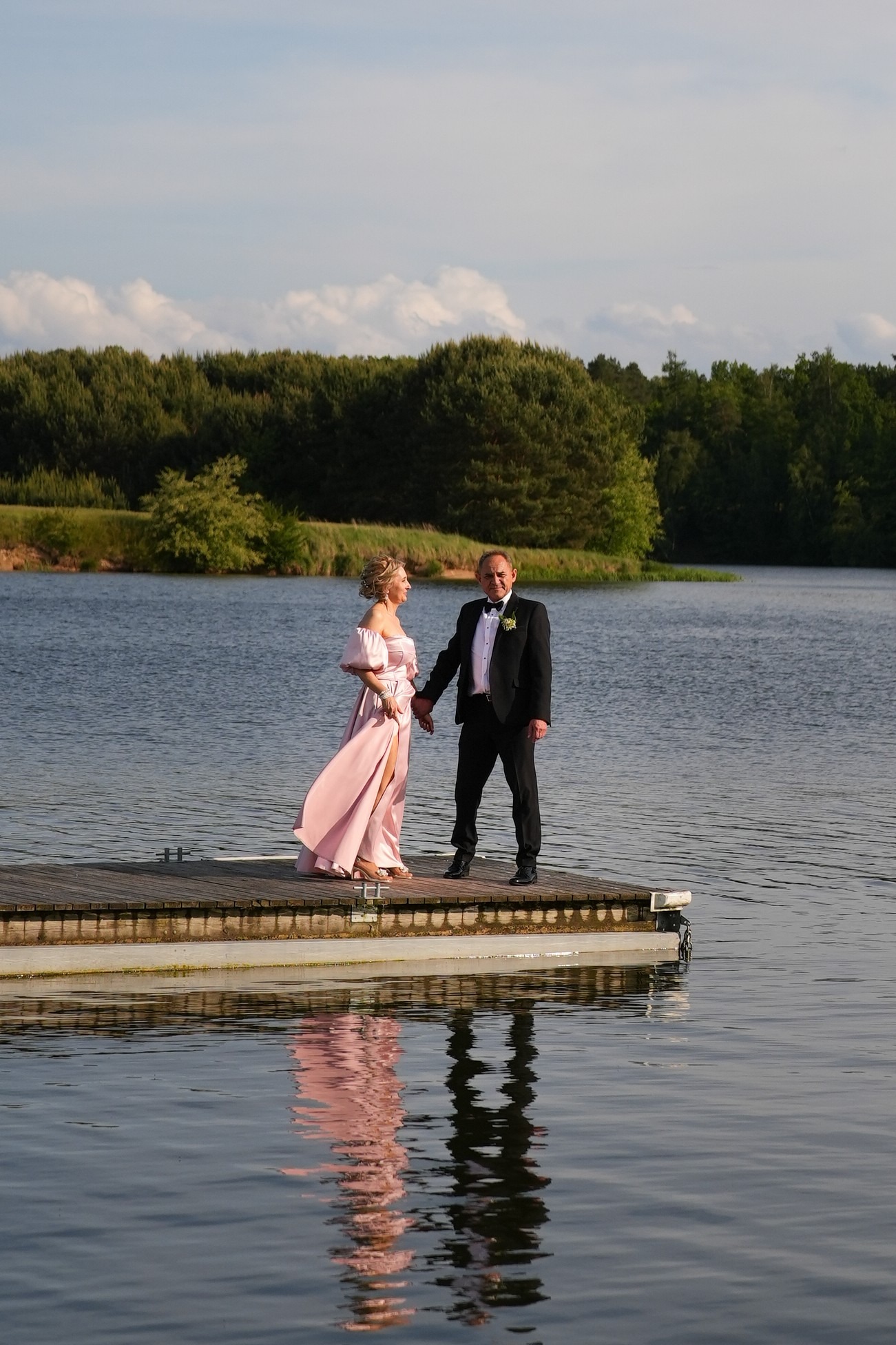 Para w eleganckich strojach stoi na drewnianym pomoście nad jeziorem, trzymając się za ręce. W tle widoczny las i błękitne niebo z chmurami.
