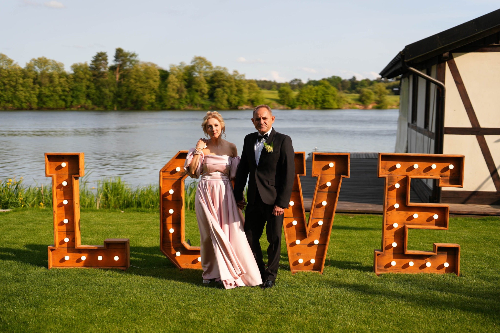 Elegancka para w strojach wieczorowych pozuje na tle jeziora z dużym napisem LOVE z żarówkami, tworząc romantyczną scenerię. Sesja plenerowa w słoneczny dzień.