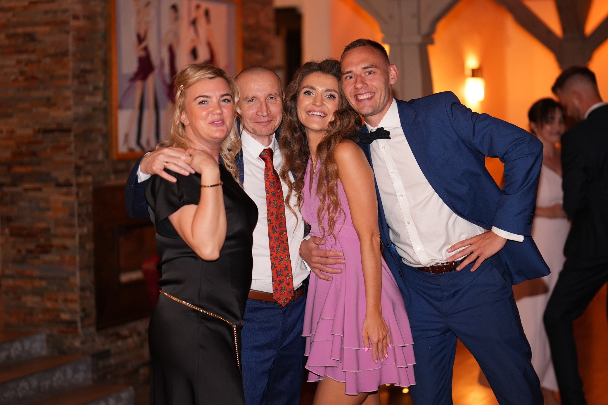Grupa uśmiechniętych osób pozuje do zdjęcia na tle ceglanej ściany i schodów. W tle widoczni inni goście i fragment dekoracji sali.