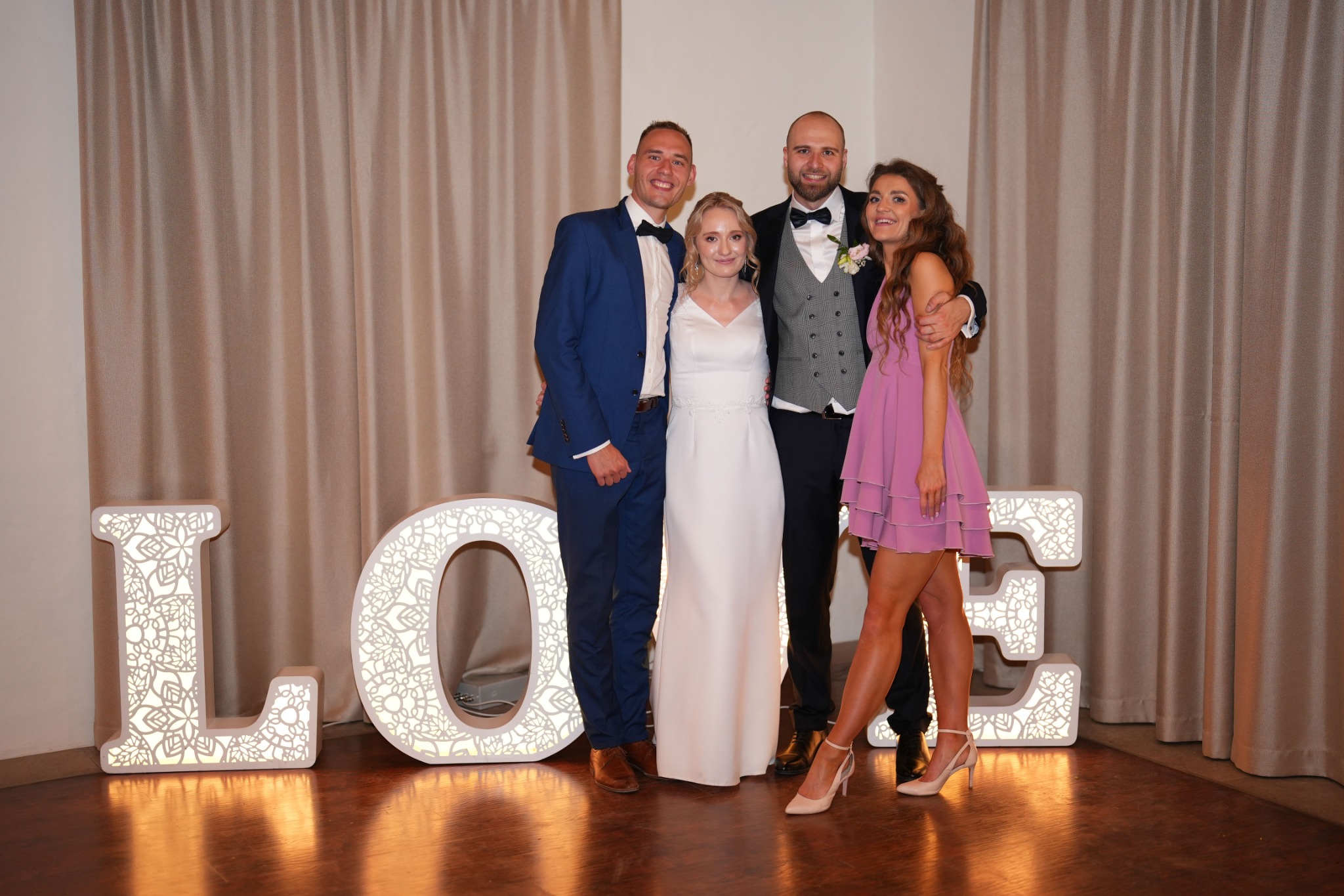 Uśmiechnięta para młoda w otoczeniu gości pozuje do zdjęcia na tle dekoracji LOVE z podświetleniem. Eleganckie stroje, ciepłe oświetlenie i beżowe zasłony w tle.