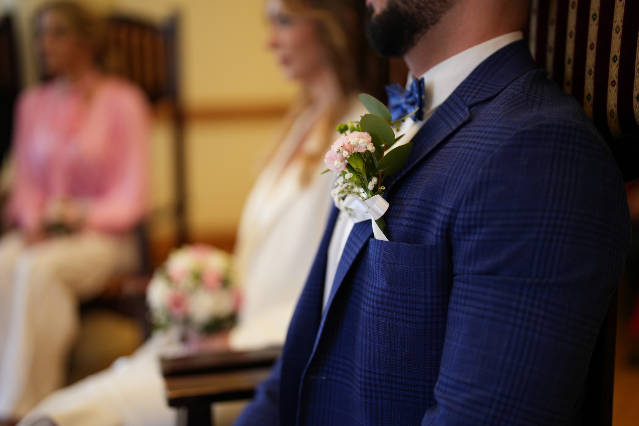 Detal męskiej stylizacji ślubnej: niebieska krata na marynarce, butonierka z różowych kwiatów i błękitna mucha. W tle rozmyta sylwetka panny młodej.