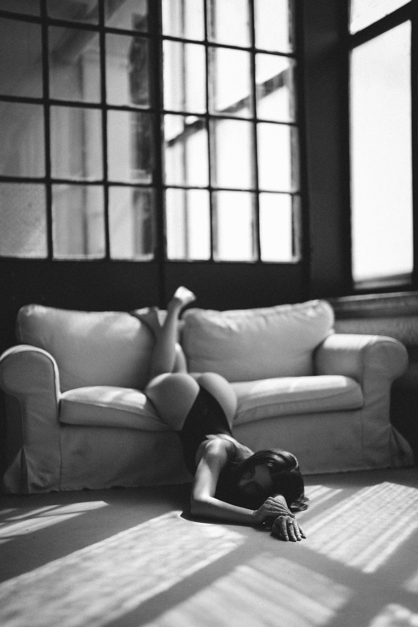 Czarno-białe zdjęcie kobiety leżącej na brzuchu na sofie w pokoju z dużymi oknami, światło pada na podłogę tworząc wyraźne cienie.