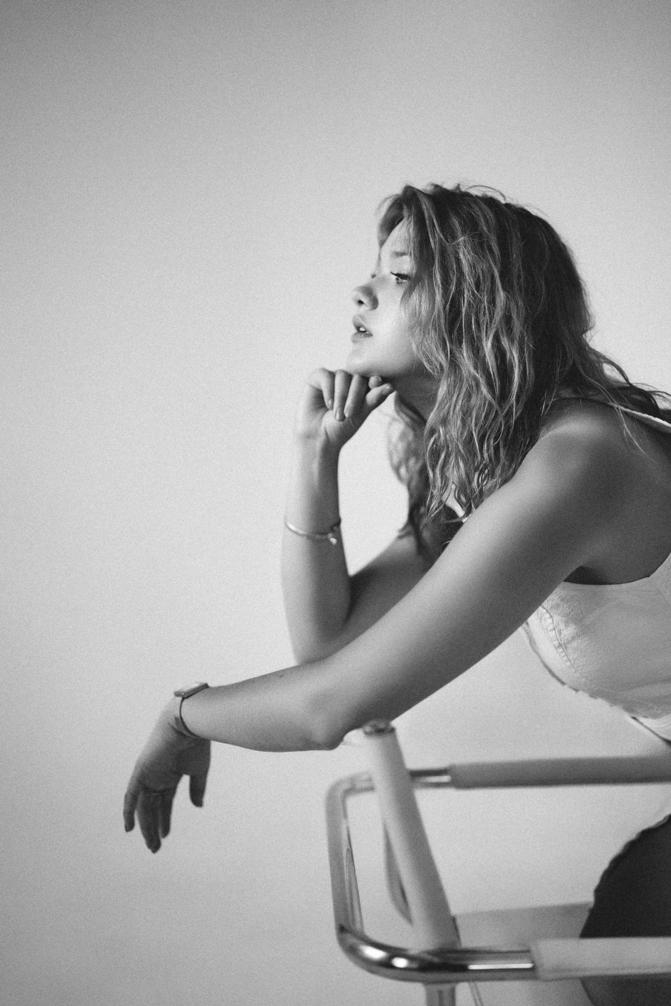 Czarno-białe ujęcie kobiety z kręconymi włosami, opierającej brodę na dłoni, zamyślona, w jasnej bluzce na tle jasnej ściany. Na ręce ma zegarek i bransoletkę.