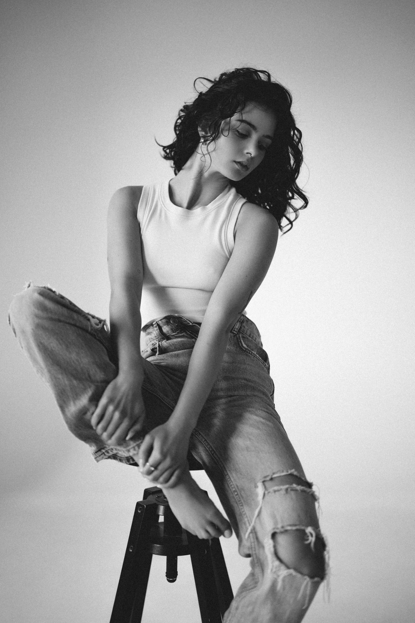 Czarno-biała fotografia studyjna: młoda kobieta w podkoszulku i podartych jeansach, siedząca na stołku, zamyślona, z delikatnym oświetleniem podkreślającym fakturę materiałów.