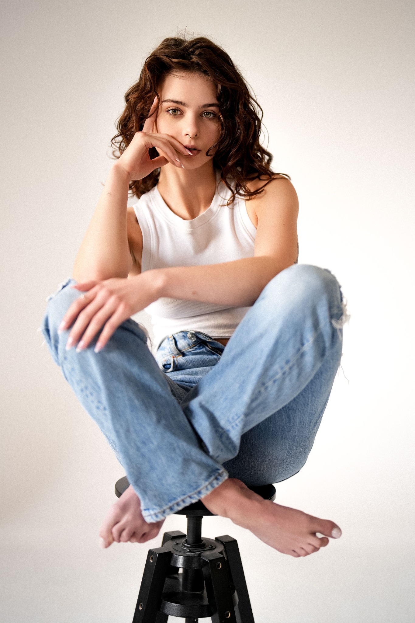 Portret kobiety w jeansach i topie, siedzącej na stołku barowym na jasnym tle. Naturalna sesja w Bydgoszczy, subtelny makijaż, swobodna poza.