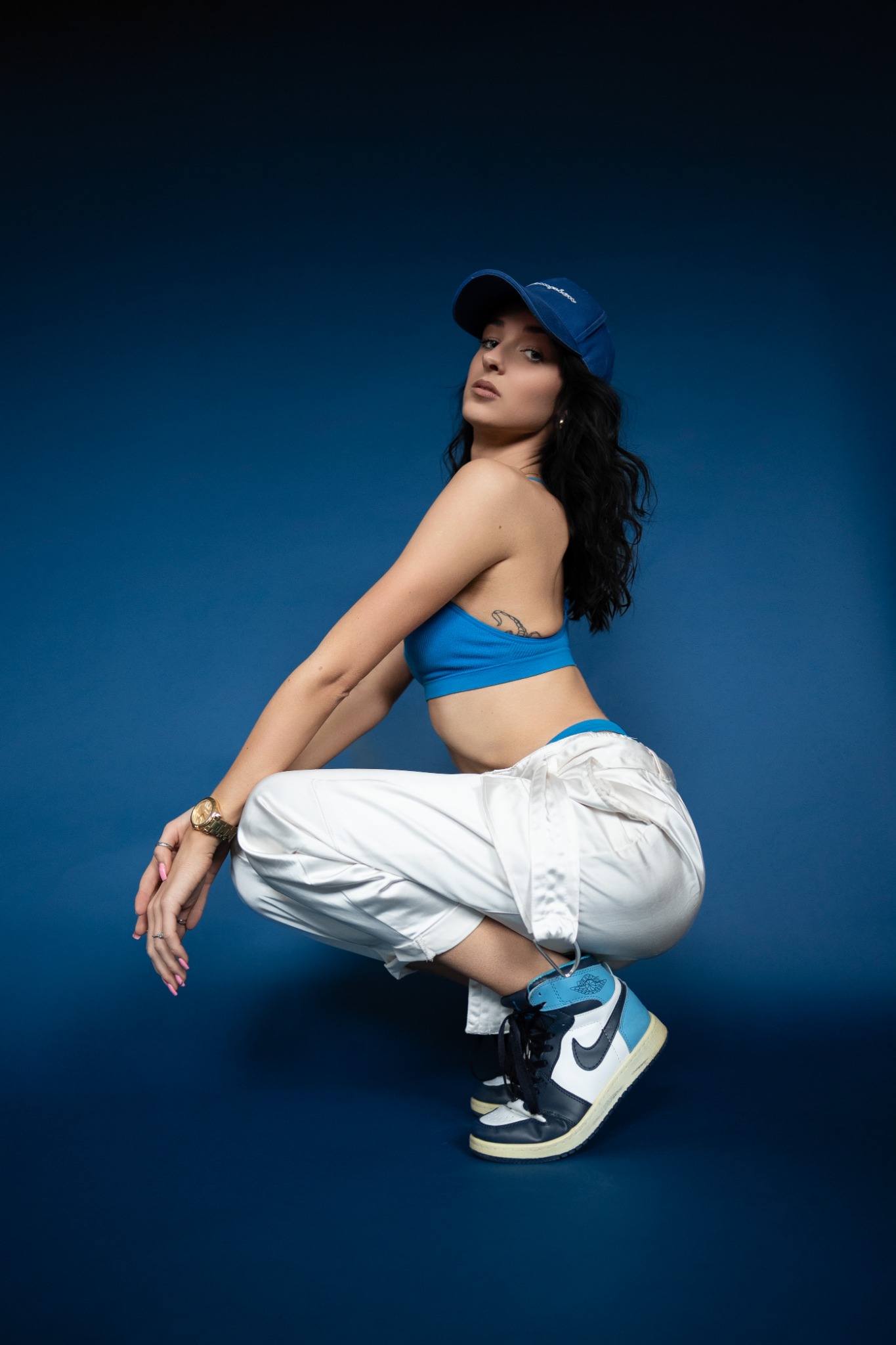 Kobieta w sportowym stroju, kucająca na tle jednolitego, ciemnoniebieskiego tła. Ubrana w błękitny top i czapkę, białe spodnie i sportowe buty.