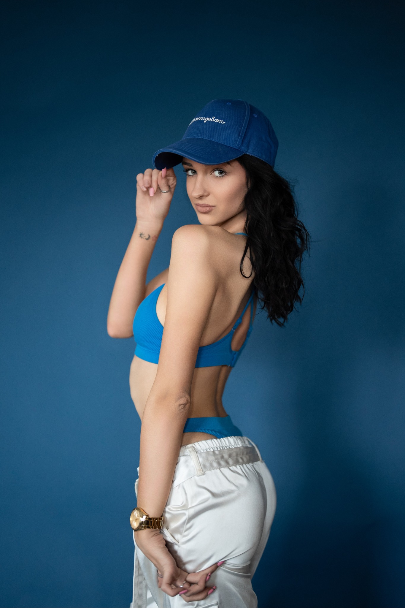Kobieta w niebieskim sportowym topie i białych spodniach, z czarnymi włosami i niebieską czapką, pozuje na jednolitym, ciemnoniebieskim tle.