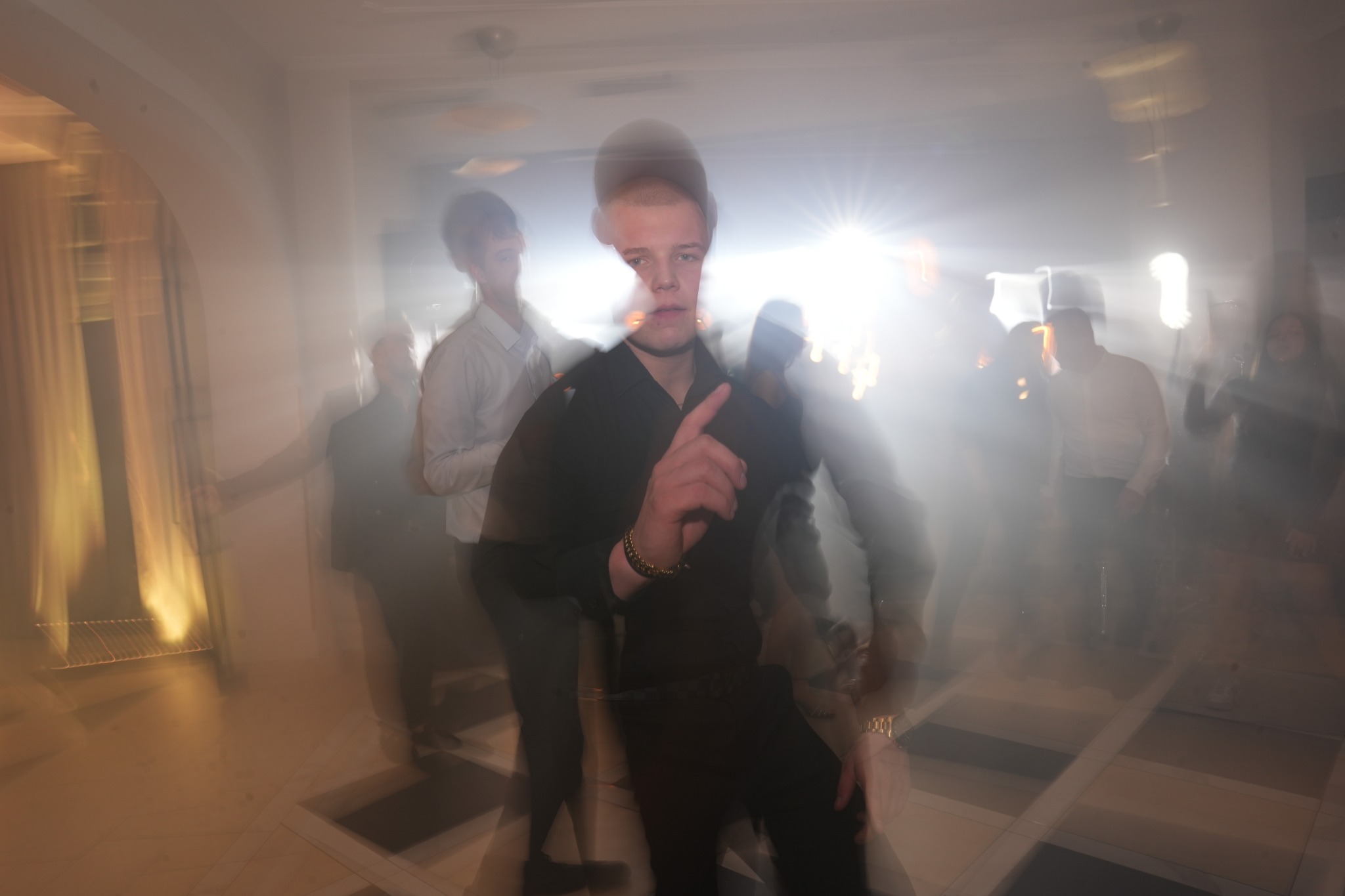 Rozmyte zdjęcie mężczyzny w czarnej koszuli na parkiecie podczas imprezy. W tle widoczni inni ludzie i oświetlenie sceniczne.