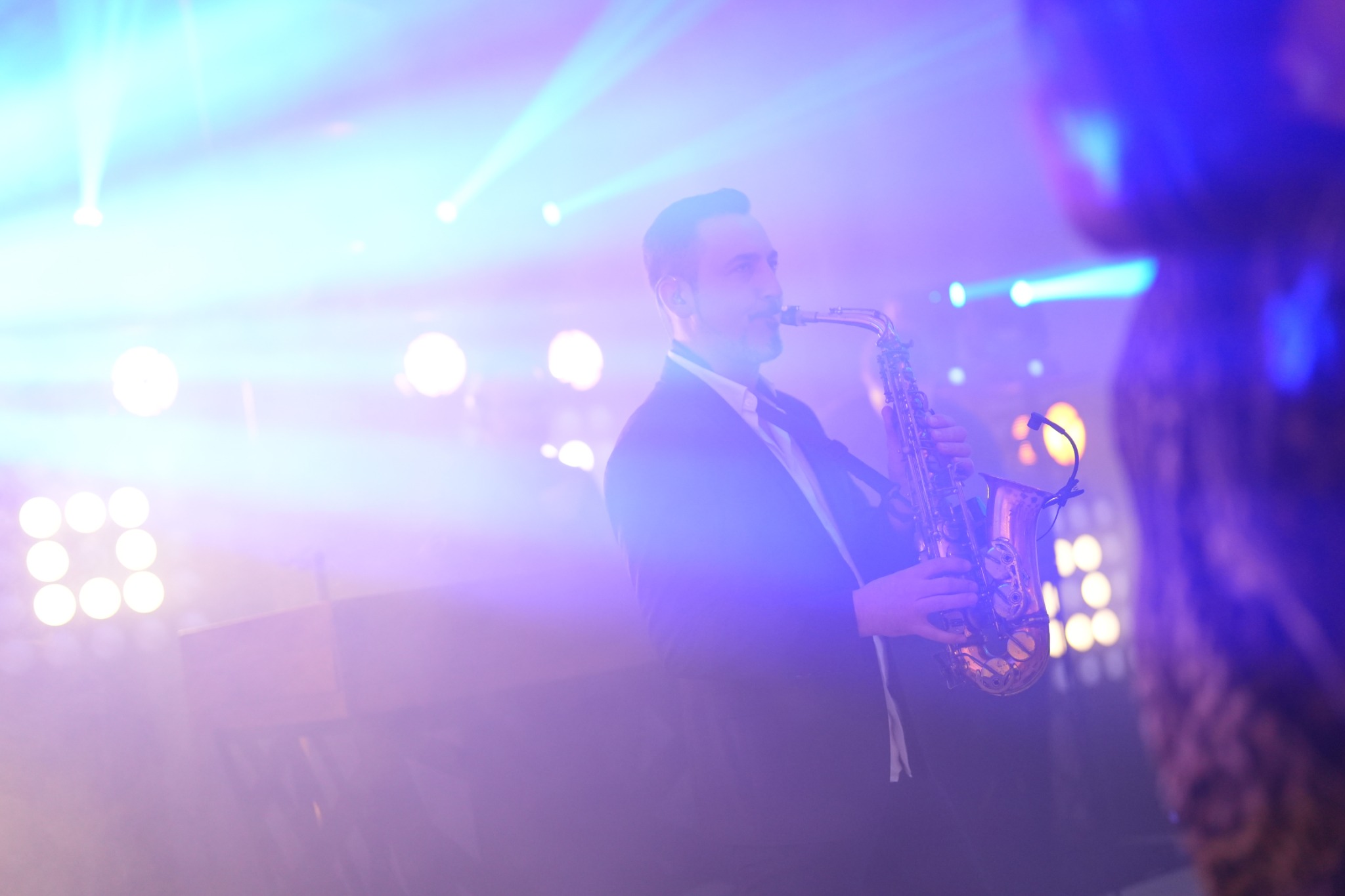 Saksofonista w trakcie występu na scenie, oświetlony reflektorami w odcieniach niebieskiego i fioletu. Widoczny z profilu, w garniturze, z instrumentem na tle świateł.