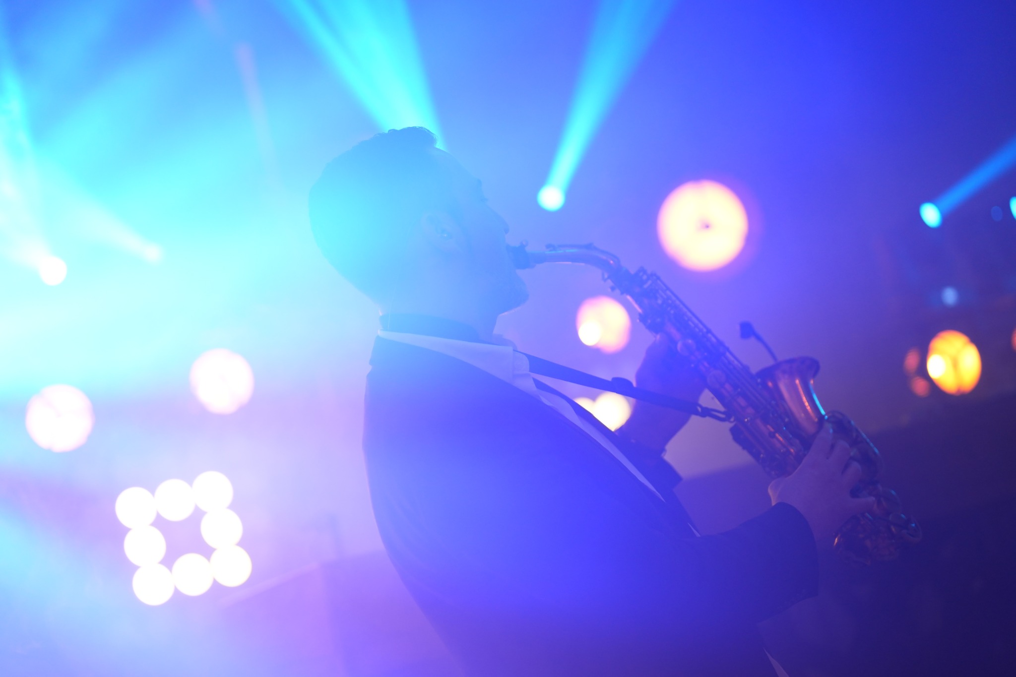 Saksofonista w garniturze gra na instrumencie w niebieskim oświetleniu scenicznym. Ujęcie z profilu, widoczne światła reflektorów w tle.