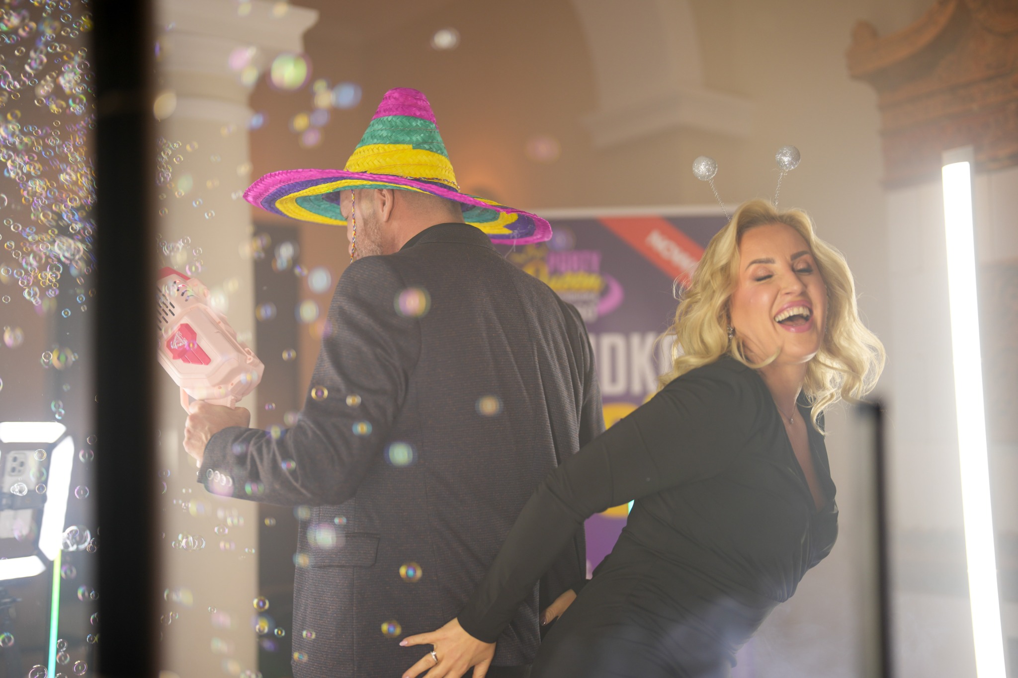 Radosna kobieta w czarnej sukience i opasce z antenkami, w otoczeniu baniek mydlanych, pozuje z mężczyzną w kapeluszu sombrero i z maszyną do baniek. Scena eventowa.