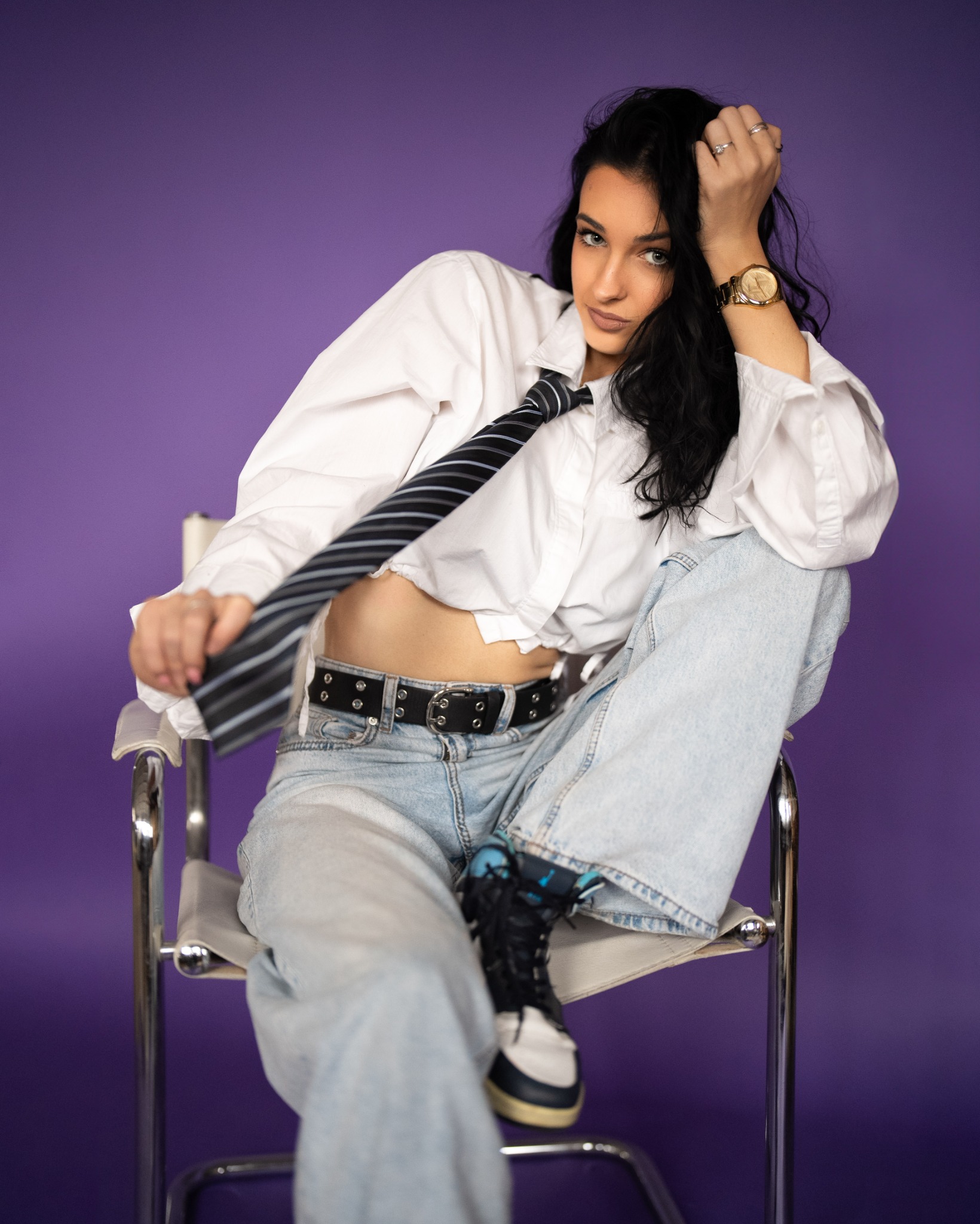 Portret kobiety w luźnej koszuli, jeansach i krawacie, siedzącej na krześle na fioletowym tle. Widoczny zegarek i buty sportowe.