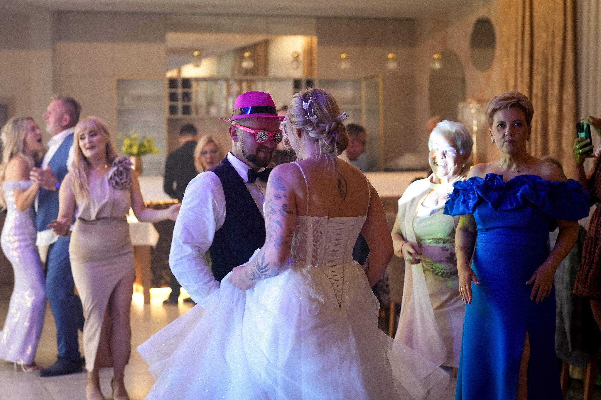Para młoda w Bydgoszczy podczas pierwszego tańca, pan młody w różowym kapeluszu i okularach. Goście weselni w tle. Ujęcie z bliska, ciepłe oświetlenie.