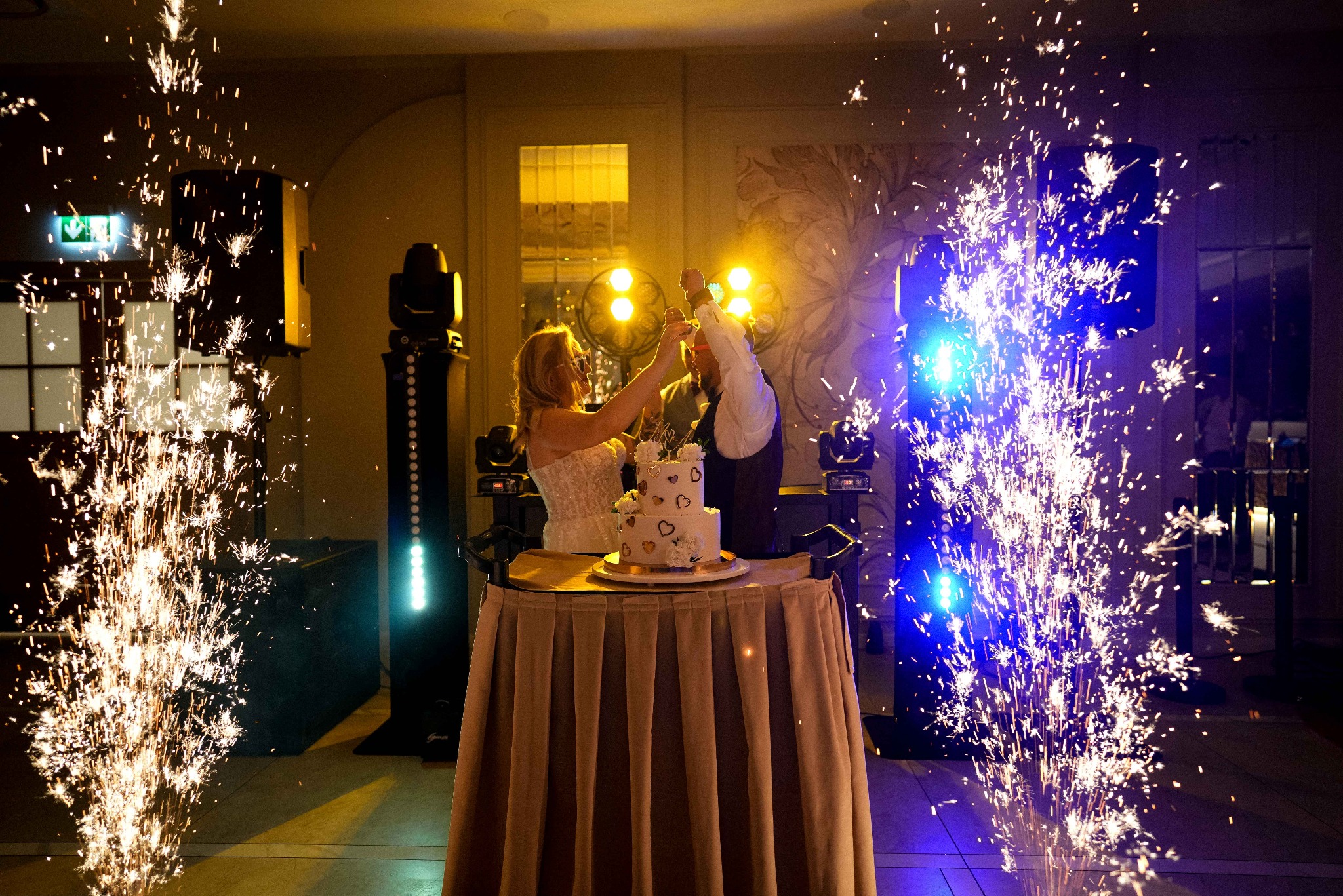 Para młoda kroi tort weselny, otoczona fontannami iskier. Dekoracje sali, oświetlenie sceniczne i dwupiętrowy tort z sercami.