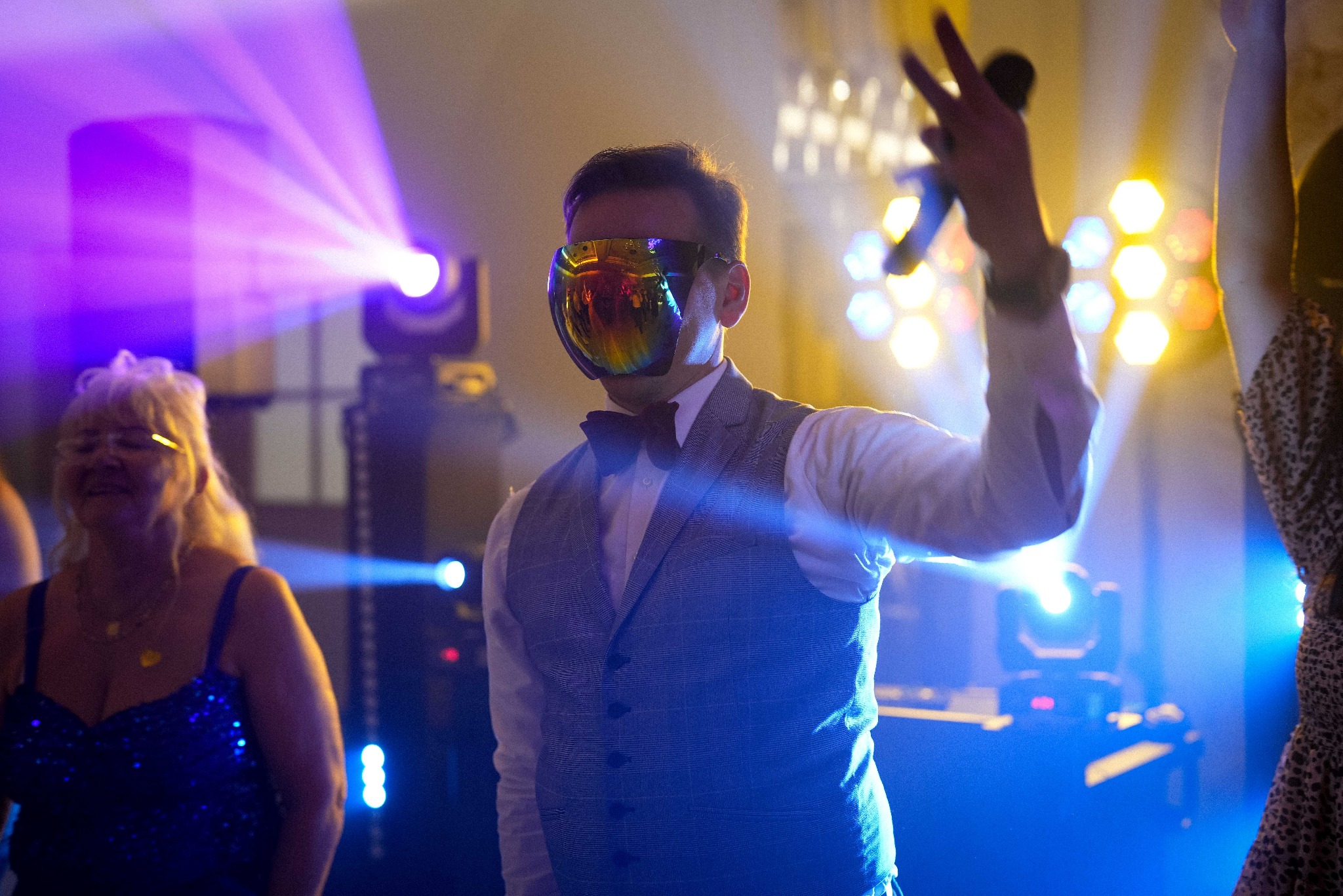 Osoba w masce na twarzy, w eleganckiej kamizelce i muszce, trzyma mikrofon na tle sceny z oświetleniem podczas wydarzenia.