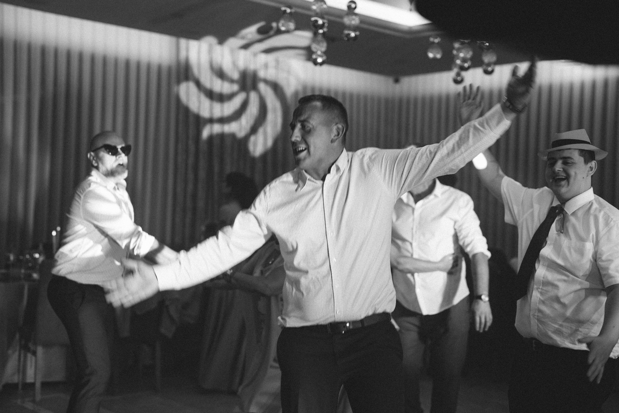 Czarno-białe zdjęcie z imprezy: mężczyźni w koszulach tańczą, jeden w kapeluszu, inny w okularach, w tle dekoracje i światła. Radosna atmosfera.