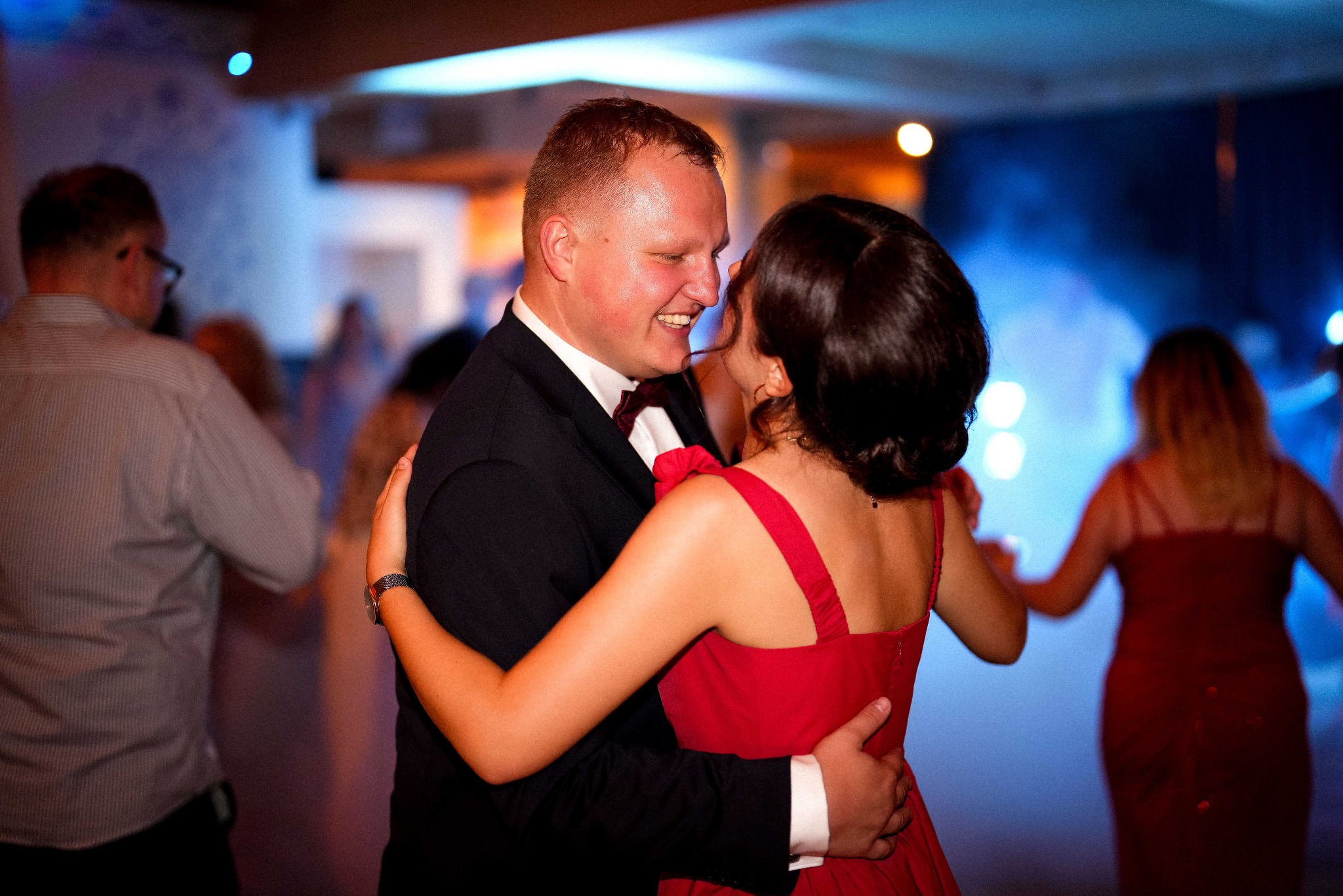 Uśmiechnięta para w tańcu, mężczyzna w smokingu z bordową muchą, kobieta w czerwonej sukience, w tle rozmyte postacie i niebieskie światła.