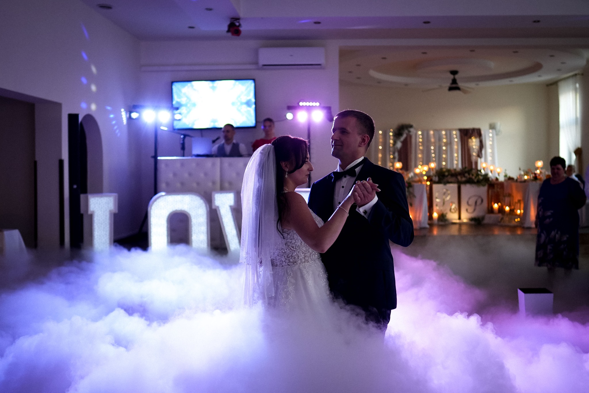 Para młoda w tańcu, otoczona sztuczną mgłą na sali weselnej z dekoracjami i napisem LOVE w tle. Romantyczna atmosfera, subtelne oświetlenie.