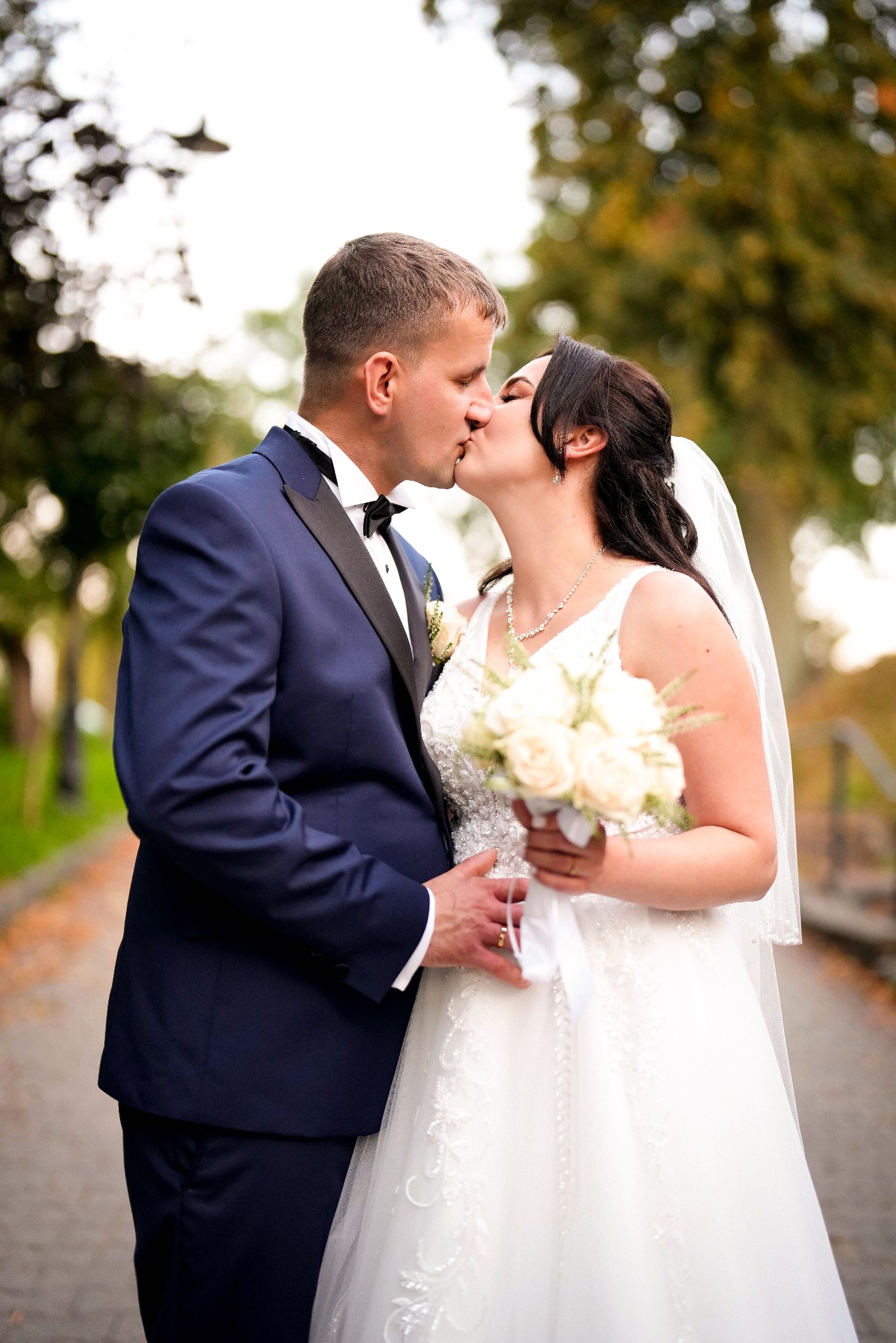 Pocałunek nowożeńców w plenerze: panna młoda w białej sukni z bukietem, pan młody w granatowym smokingu. Romantyczna chwila na tle jesiennych drzew.