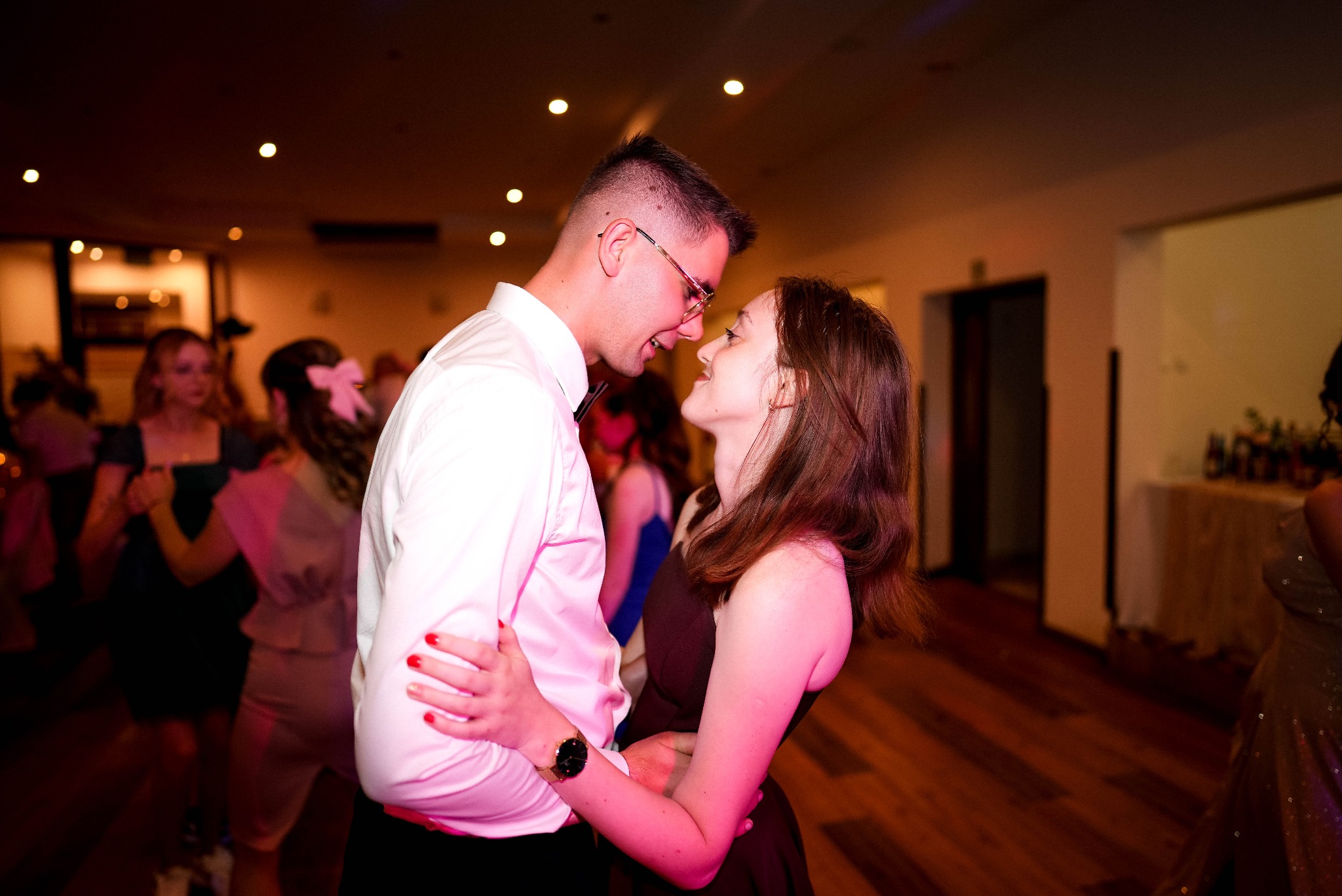 Para w objęciach na parkiecie, mężczyzna w okularach i koszuli, kobieta w sukience, ciepłe światło, w tle tańczący goście, parkiet z drewna.