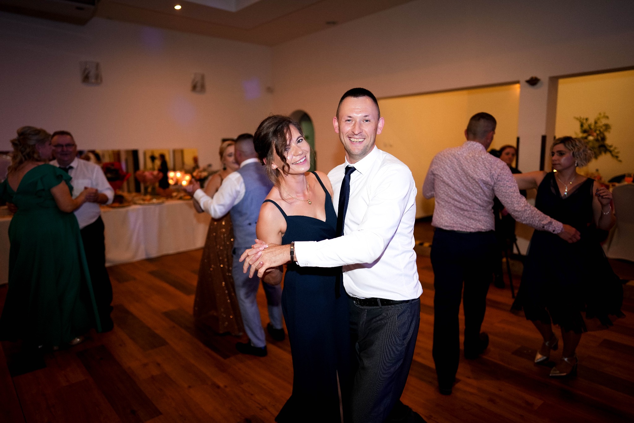 Uśmiechnięta para w tańcu na parkiecie podczas uroczystości, w tle inni goście i bufet. Mężczyzna w białej koszuli, kobieta w granatowej sukience.