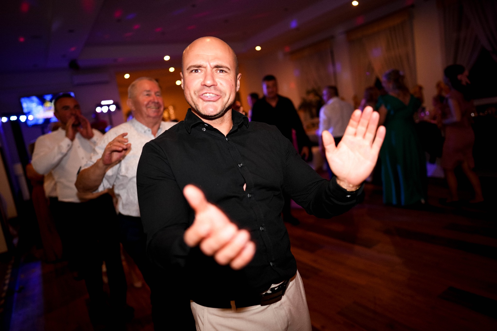 Mężczyzna w czarnej koszuli tańczy na parkiecie podczas imprezy, w tle rozmazane sylwetki gości i dekoracje świetlne.