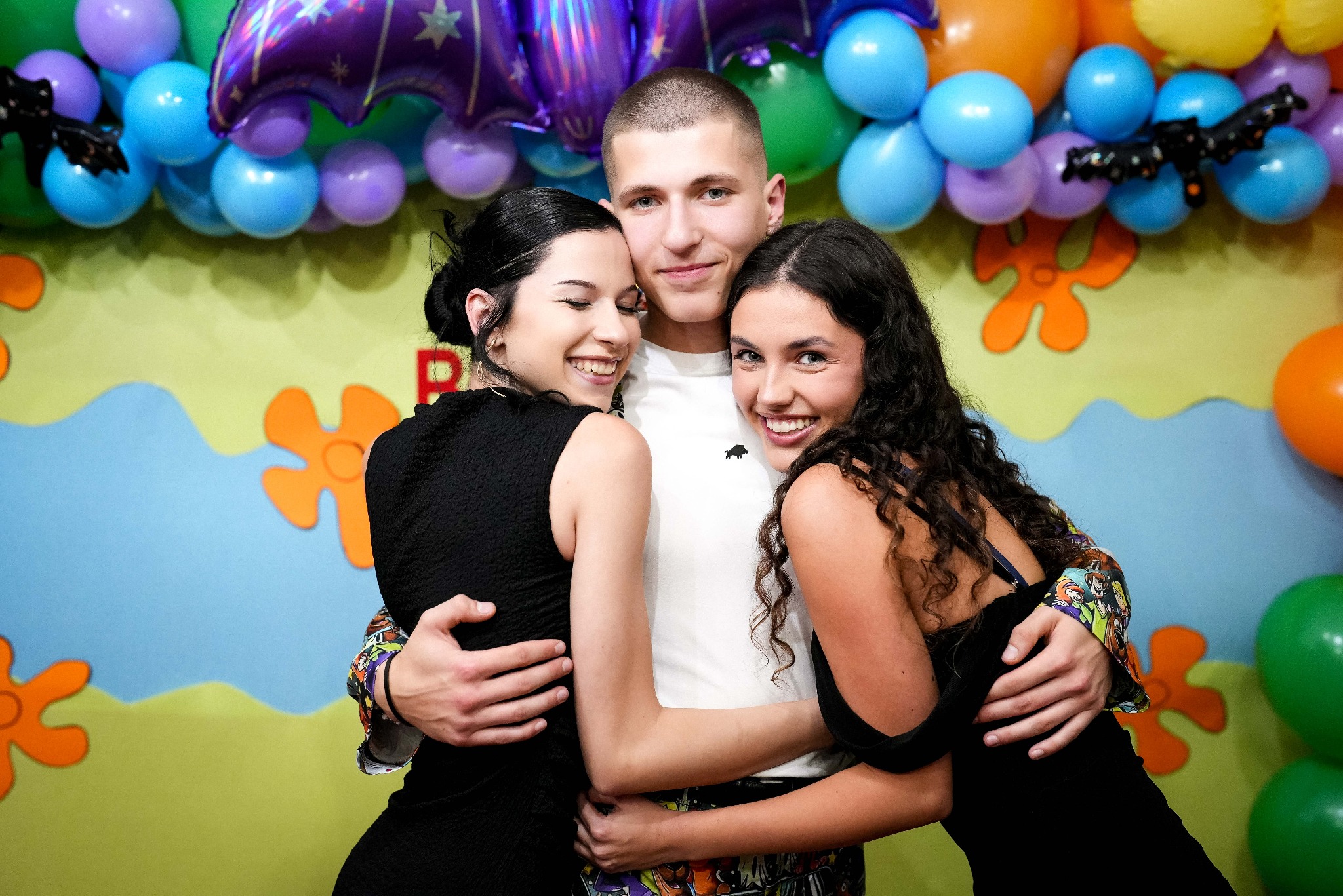 Uśmiechnięty mężczyzna w objęciach dwóch kobiet na tle kolorowych balonów i dekoracji ściennych. Radosna atmosfera, bliskie relacje, imprezowy klimat.