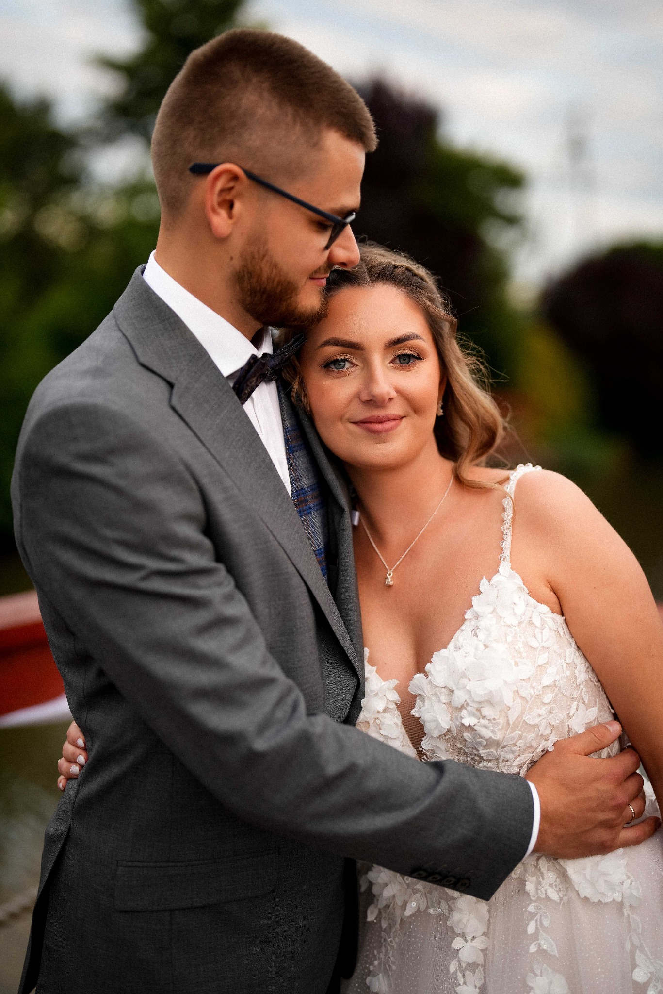 Uśmiechnięta panna młoda w koronkowej sukni, oparta o ramię pana młodego w szarym garniturze z muchą, na tle zieleni i rozmytego nieba.