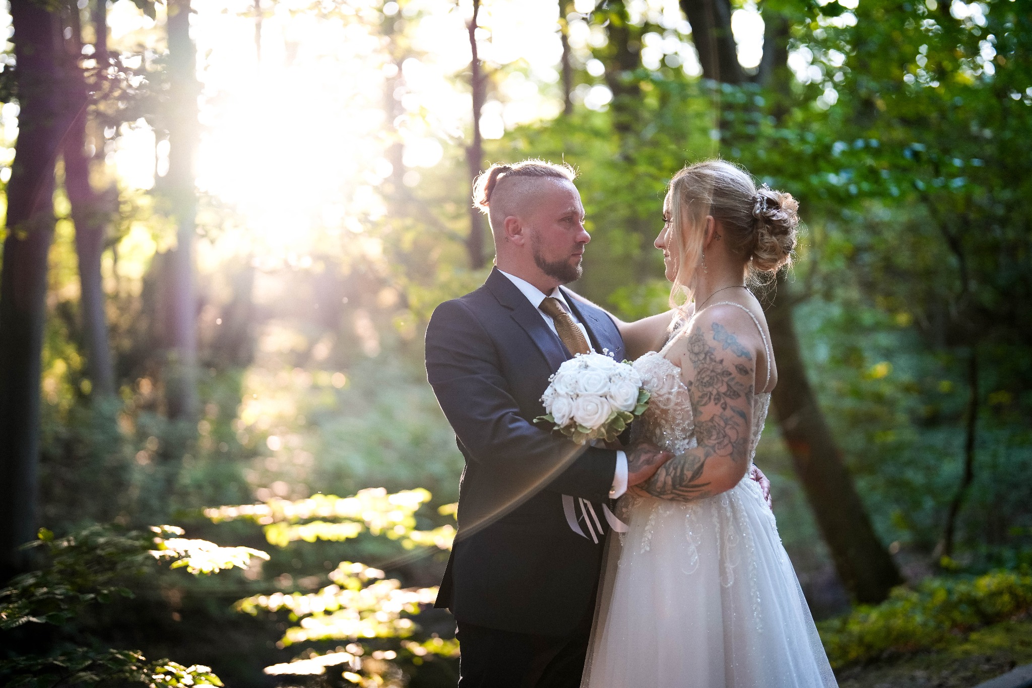 Para młoda w leśnej scenerii, on w ciemnym garniturze z bukietem, ona w białej sukni z tatuażami, delikatne światło słoneczne w tle, romantyczny kadr.