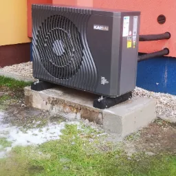 Czarna jednostka zewnętrzna klimatyzacji marki Kaisai, zamontowana na betonowym fundamencie przy ścianie budynku z widocznymi przewodami chłodniczymi i fragmentami trawnika z resztkami śniegu.