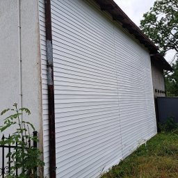 Blask Time - Boczny widok wyczyszczonej, białej elewacji domu z sidingu, z widocznym fragmentem rynny i roślinnością przy ścianie. Ujęcie z dołu, w tle drzewa i niebo.