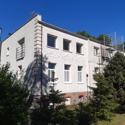 Blask Time - Dwupiętrowy, jasny budynek z elementami dekoracyjnymi, otoczony zielenią. Widoczne rusztowania przy ścianie, sugerujące prace remontowe lub czyszczenie elewacji.