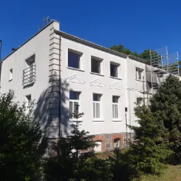 Dwupiętrowy, jasny budynek z elementami dekoracyjnymi, otoczony zielenią. Widoczne rusztowania przy ścianie, sugerujące prace remontowe lub czyszczenie elewacji.